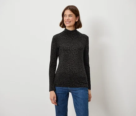 Une femme aux cheveux bruns pose en souriant dans un haut noir à manches longues et un jean bleu.