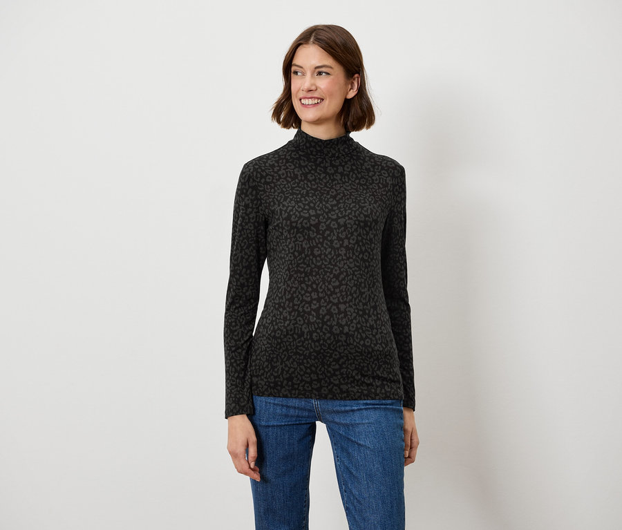 Une femme aux cheveux bruns pose en souriant dans un haut noir à manches longues et un jean bleu.