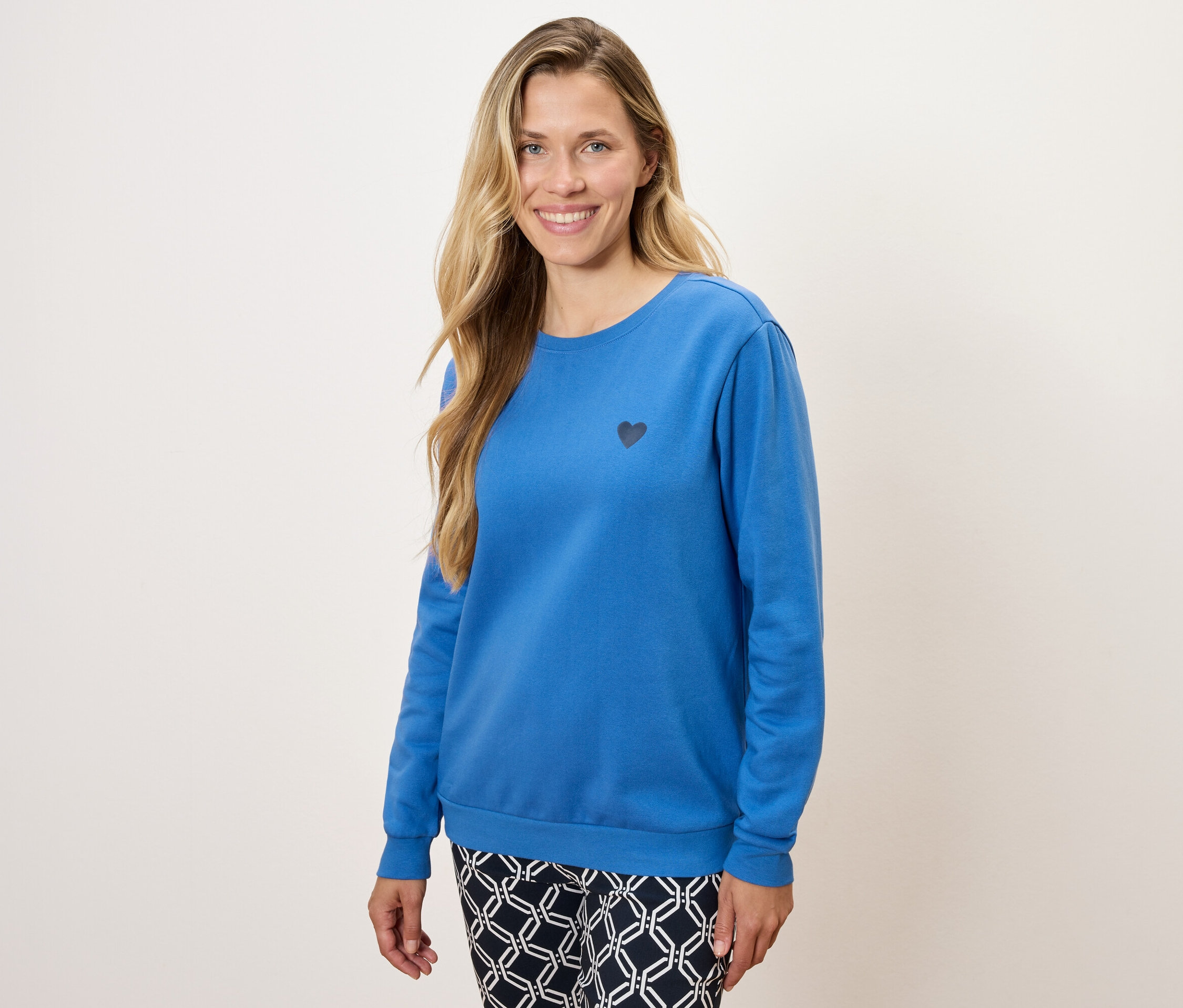 Une femme porte un pull bleu à manches longues avec un petit cœur noir et un pantalon à motifs.