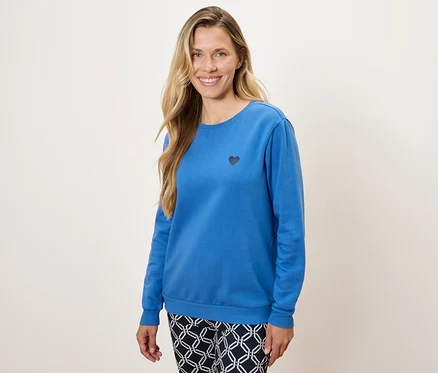 Une femme porte un pull bleu à manches longues avec un petit cœur noir et un pantalon à motifs.