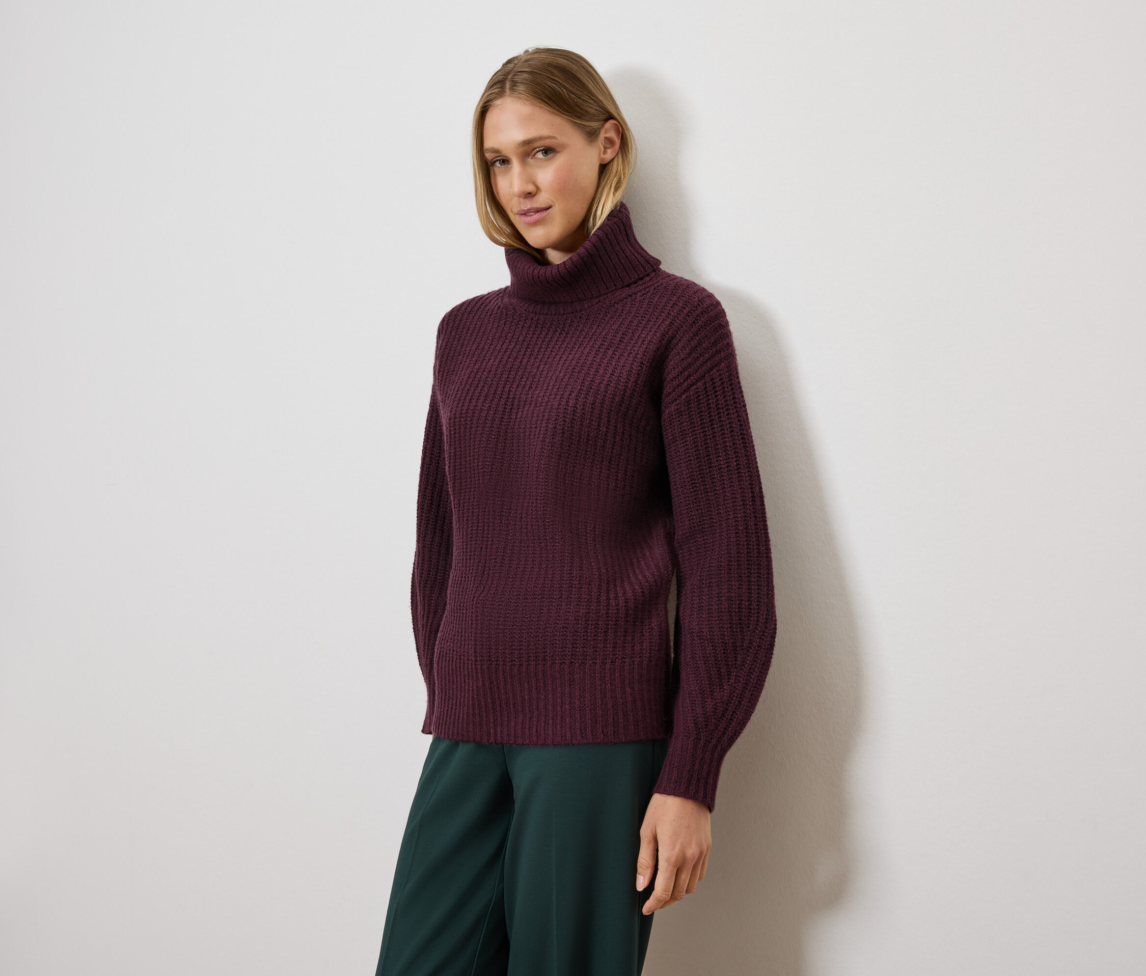 Femme posant portant un pull à col roulé violet et un pantalon vert.