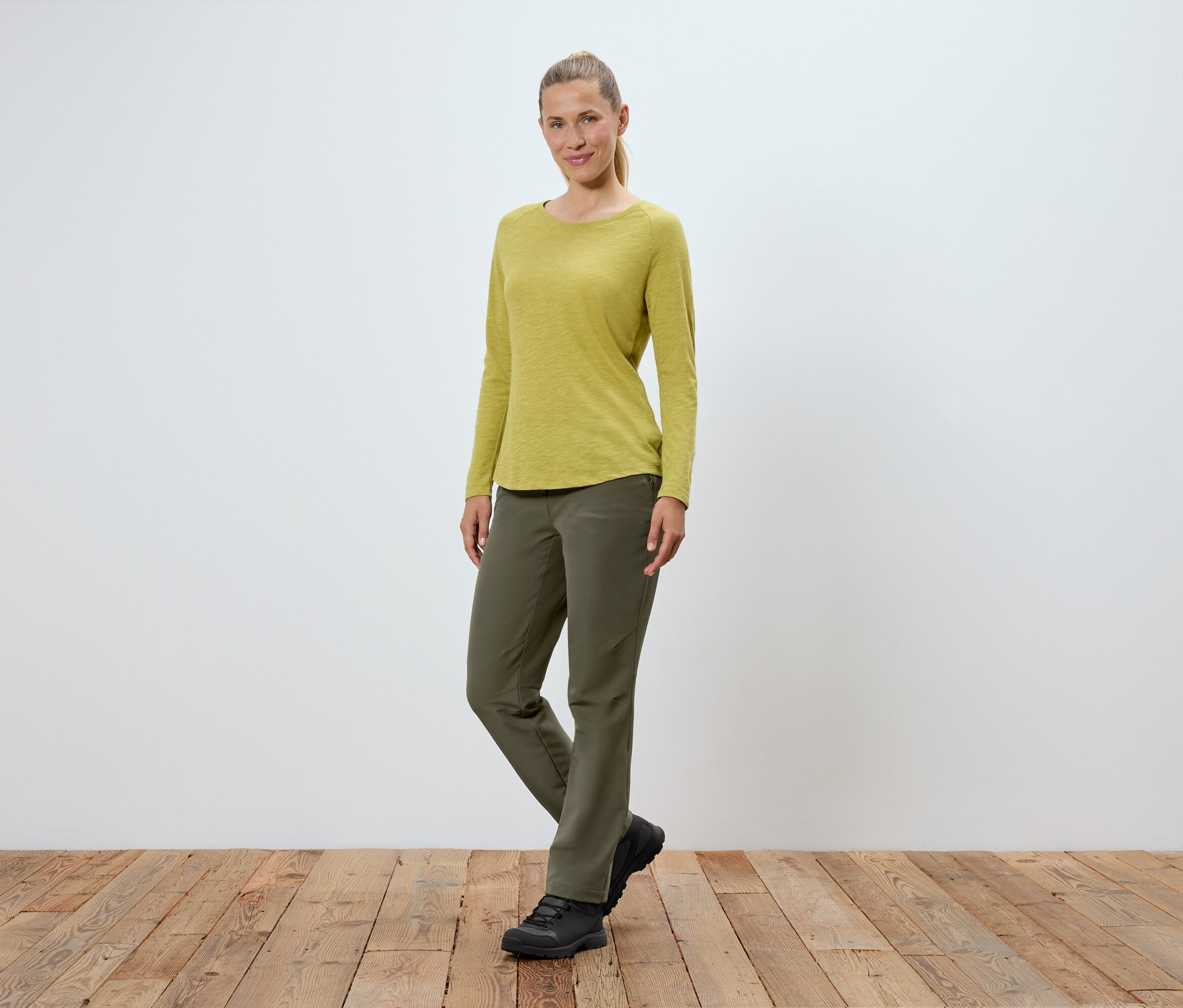 Une femme portant un pantalon vert olive, un t-shirt à manches longues jaune-vert et des chaussures noires se tient sur un plancher en bois devant un mur blanc.