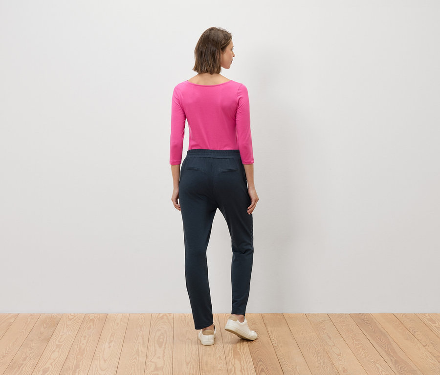 Une femme se tient dos à la caméra, elle porte un chemisier rose et un pantalon bleu.