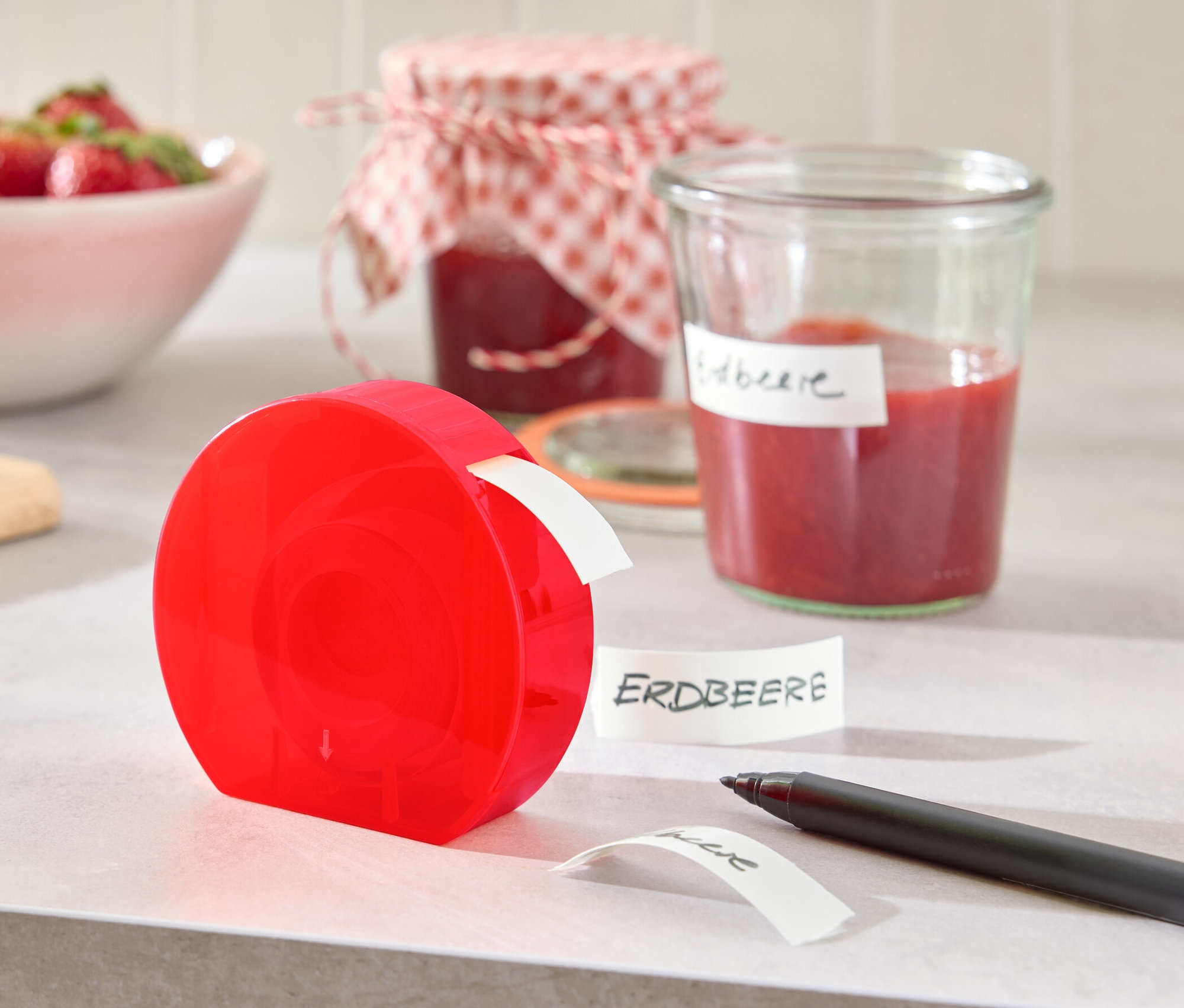 Un distributeur d'étiquettes rouge est posé sur une table à côté d'un pot de confiture et d'un bol de fraises.