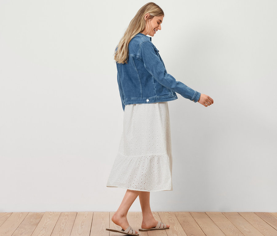 Femme marchant et portant une veste en jean, une robe avec broderie ajourée et un cabas aspect raphia.