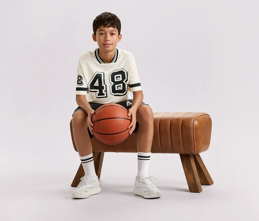 Un garçon est assis sur un banc marron et tient un ballon de basket.