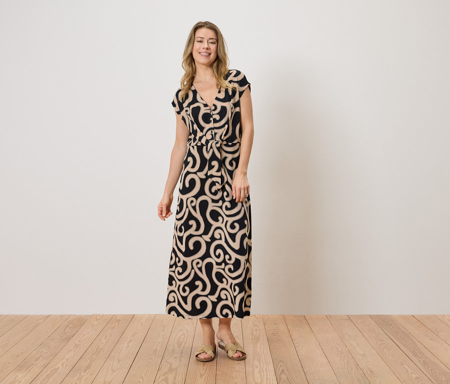 Une femme porte une longue robe à motifs avec une ceinture et des sandales plates devant un mur blanc.