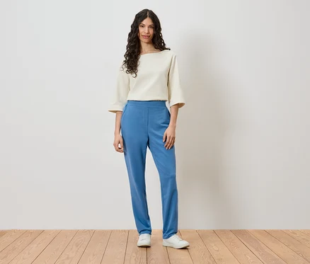 Une femme se tient devant un mur et porte un haut crème, un pantalon bleu et des chaussures blanches.