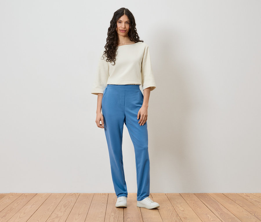 Une femme se tient devant un mur et porte un haut crème, un pantalon bleu et des chaussures blanches.