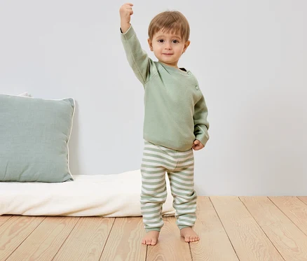 Un jeune enfant portant un pull vert clair et un pantalon rayé se tient pieds nus sur un plancher en bois et lève le poing.