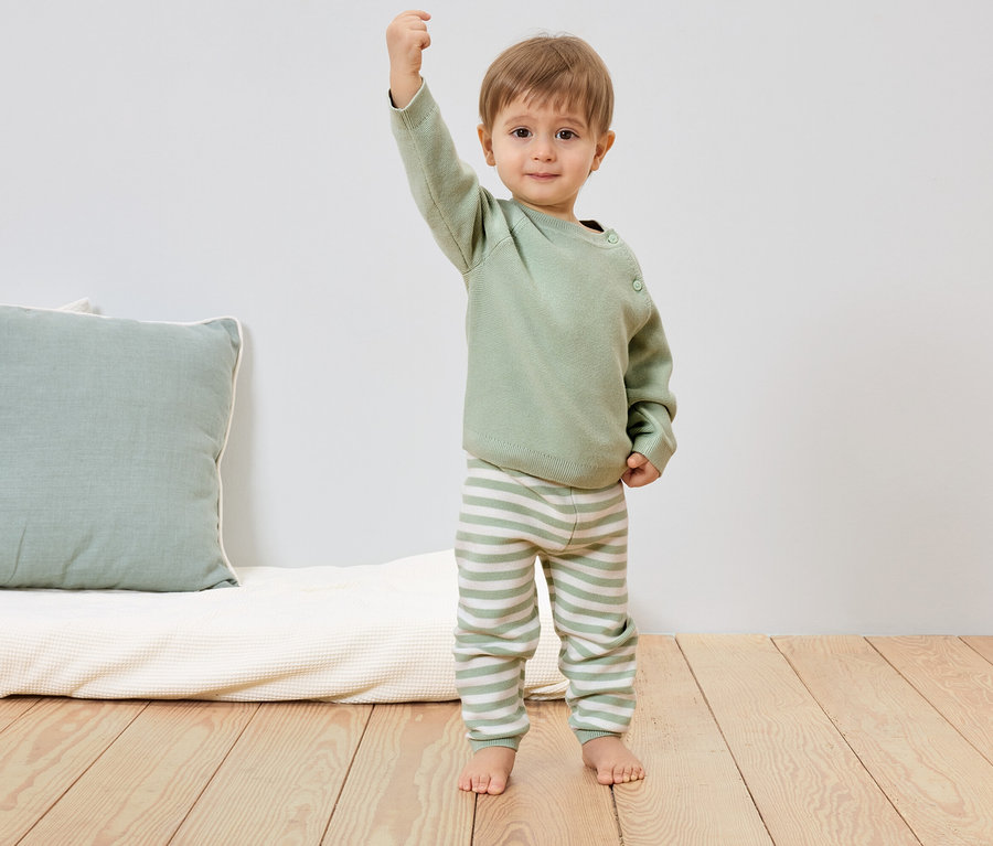 Un jeune enfant portant un pull vert clair et un pantalon rayé se tient pieds nus sur un plancher en bois et lève le poing.