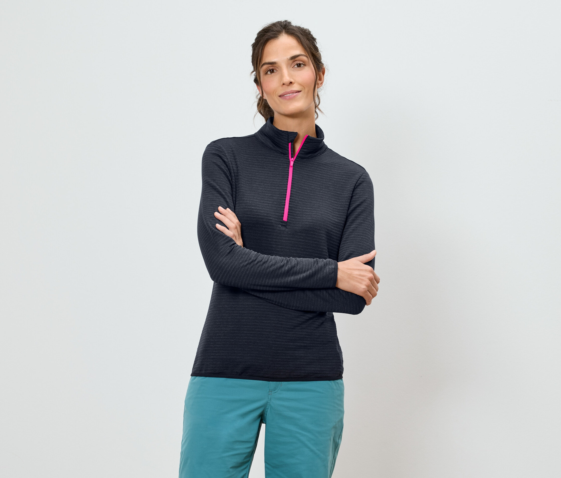 Une femme aux bras croisés porte une chemise bleu marine à manches longues avec une fermeture éclair rose et un pantalon turquoise sur un fond blanc.