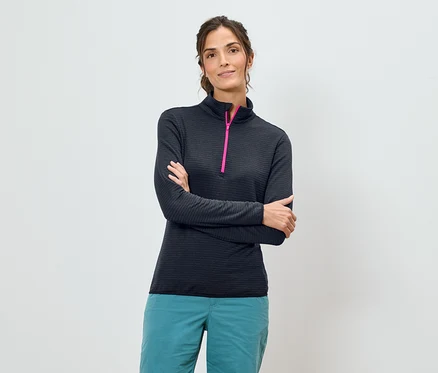 Une femme aux bras croisés porte une chemise bleu marine à manches longues avec une fermeture éclair rose et un pantalon turquoise sur un fond blanc.