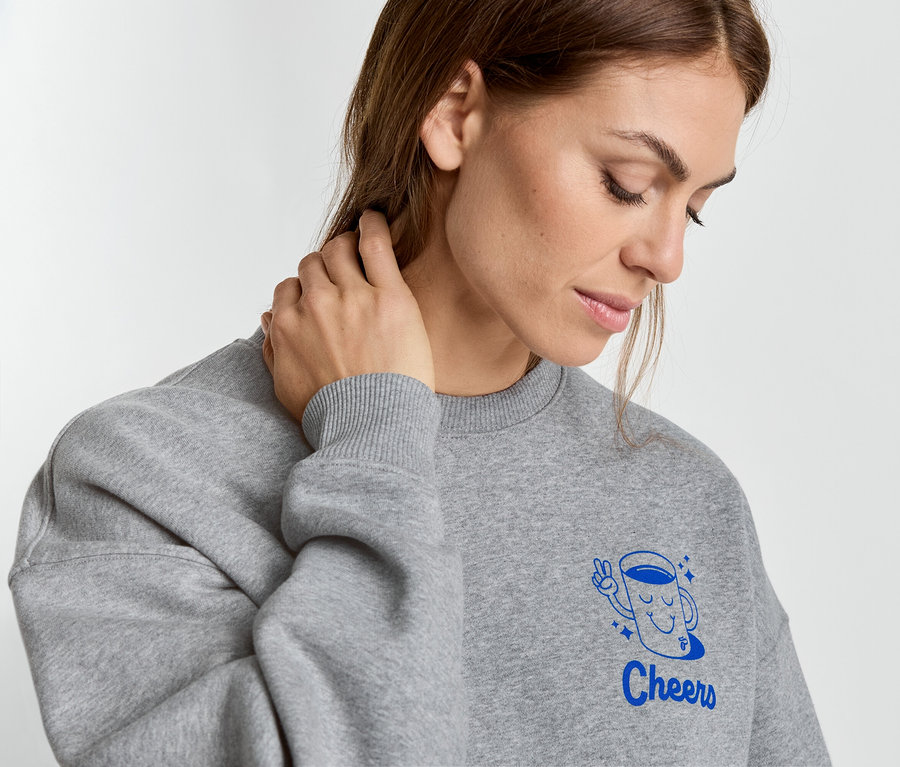 Femme portant un sweat-shirt gris avec une tasse bleue et l'inscription « Cheers » touchant son cou.