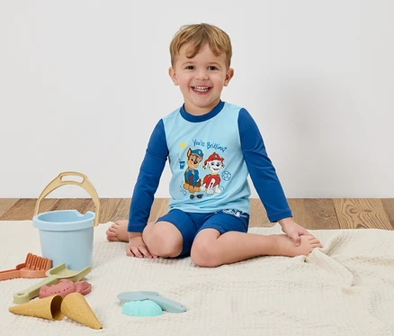 Un garçon souriant est assis sur une couverture, portant un t-shirt bleu clair Pat' Patrouille et un short bleu, entouré de jouets de plage.