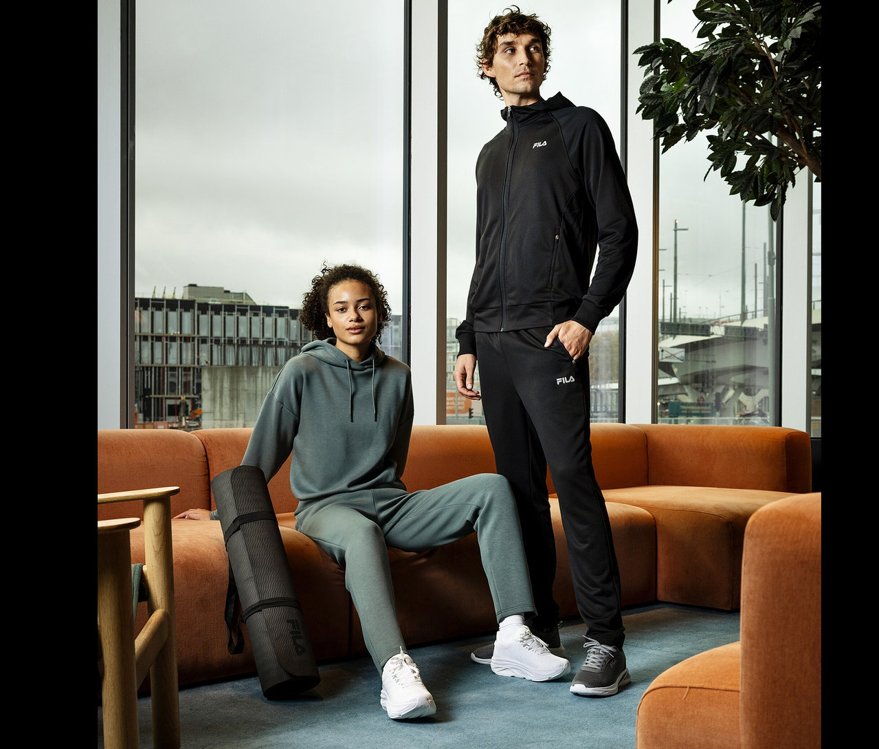 Une femme est assise sur un canapé orange avec un tapis de yoga noir. Un homme se tient à côté d'elle. Ils portent tous les deux des vêtements de sport Fila.