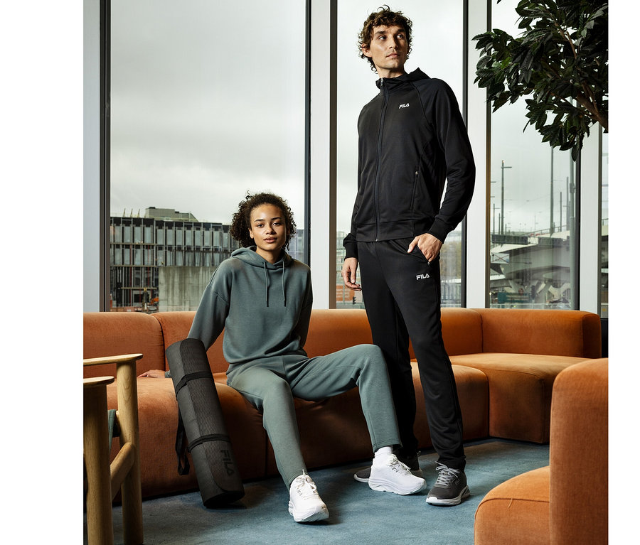 Une femme est assise sur un canapé orange à côté d'un homme debout. Elle porte un sweat à capuche et un pantalon de survêtement verts, avec un tapis de yoga noir à côté d'elle. Il porte un sweat à capuche noir et un pantalon sweat technique pour homme FILA noir.