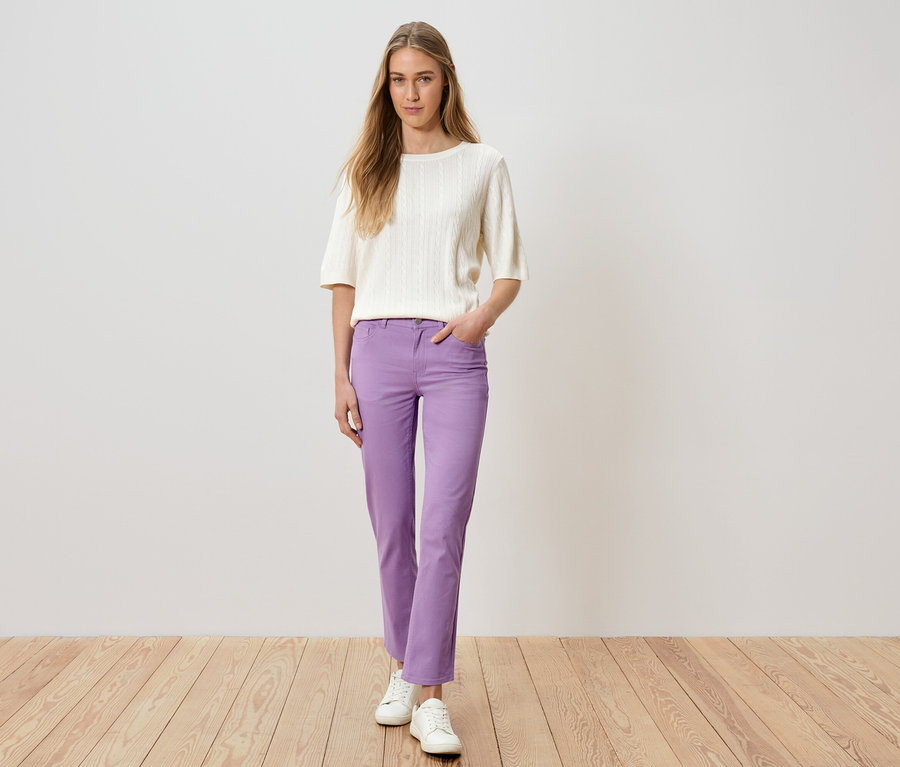 Une femme se tient debout, la main dans la poche, portant un pantalon violet, un pull blanc et des chaussures blanches.