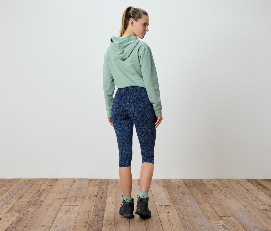 Une femme se tient dos à la caméra, portant un sweat à capuche vert menthe et un legging bleu marine à motifs floraux.