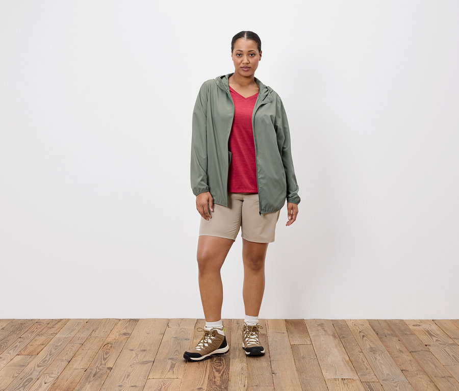 Une femme se tient devant un mur blanc, portant une veste verte, un t-shirt rouge, un short beige et des chaussures de randonnée.