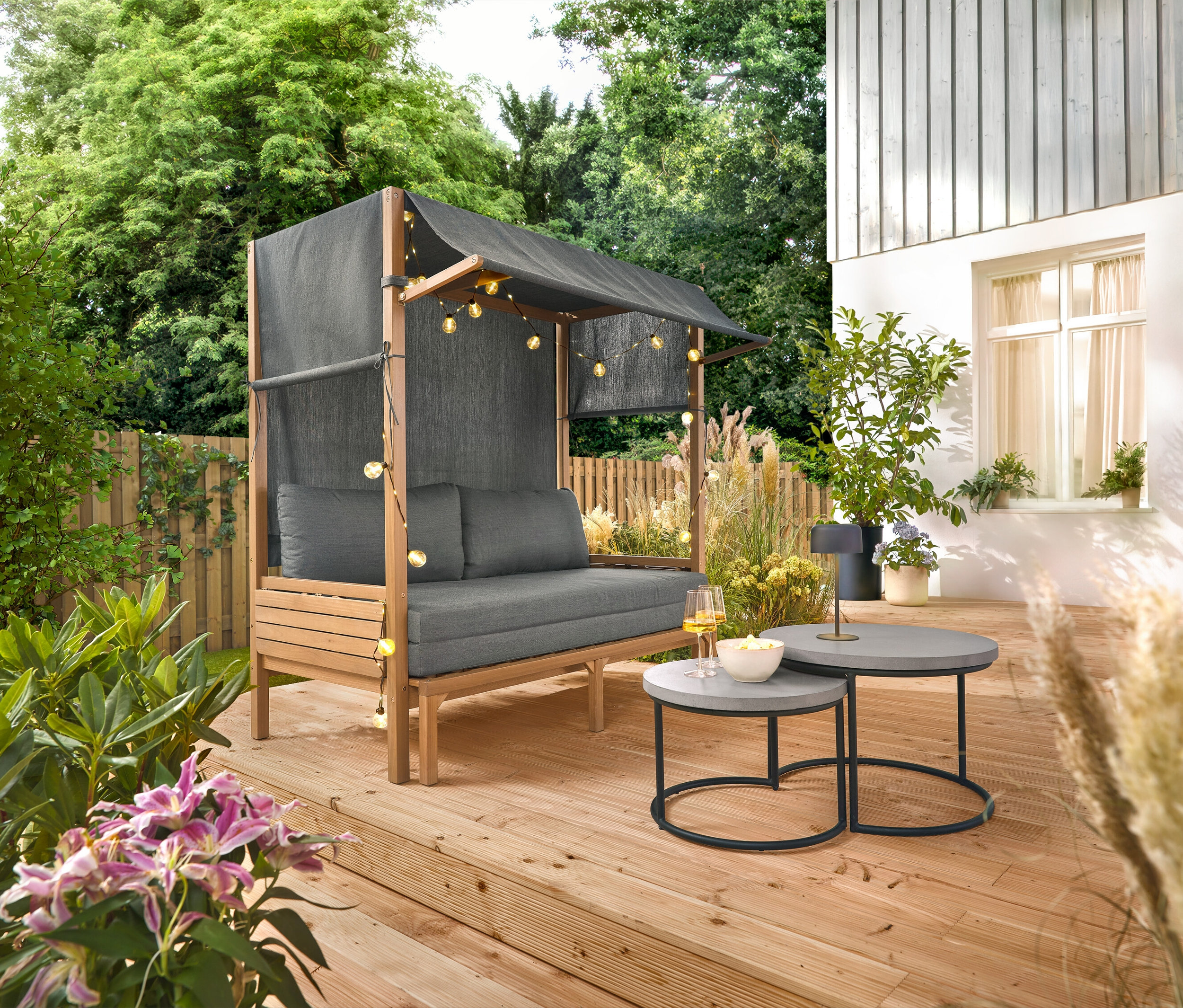 Un salon de jardin gris avec un auvent est installé sur une terrasse en bois, à côté de deux tables rondes, des boissons et des collations sont posées sur une table.