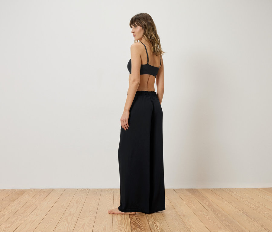 Femme de dos en soutien-gorge noir et pantalon ample devant un mur blanc.
