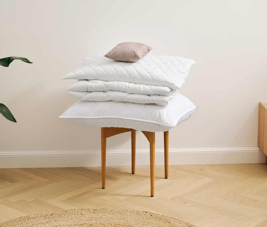 Un oreiller lavable à haute température (env. 80 x 40) repose sur une couette lavable à haute température de taille normale, empilés sur un tabouret.