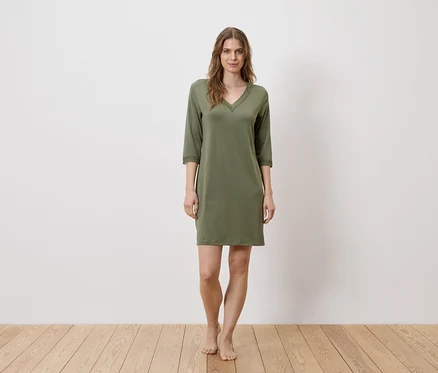 Une femme se tient pieds nus devant un mur blanc, portant une chemise de nuit verte.