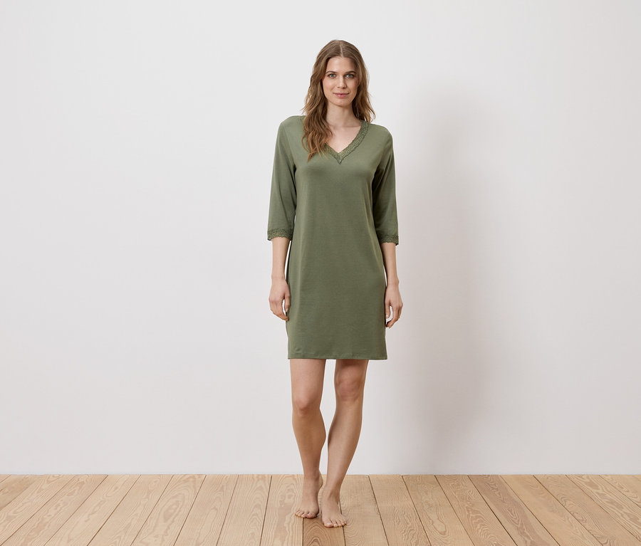 Une femme se tient pieds nus devant un mur blanc, portant une chemise de nuit verte.