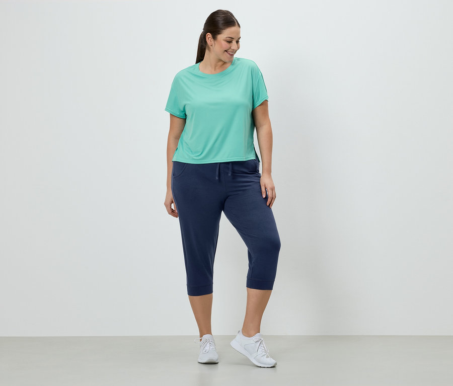 Une femme portant un t-shirt turquoise, un pantacourt bleu marine et des baskets blanches se tient devant un mur blanc.