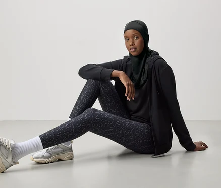 Une femme portant un hijab noir, un survêtement et des baskets argentées est assise par terre.