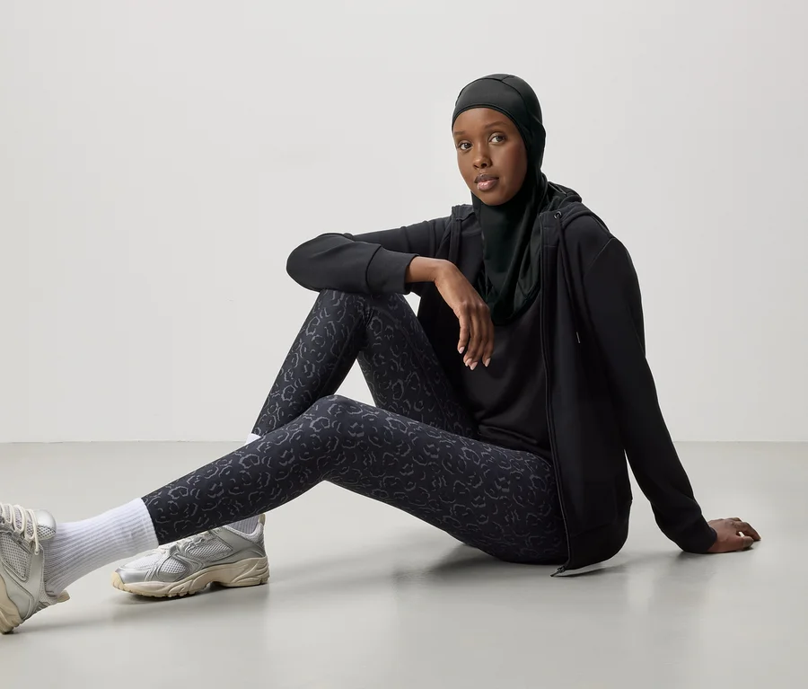Une femme portant un hijab noir, un survêtement et des baskets argentées est assise par terre.