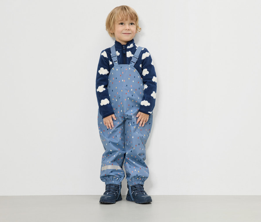 Un petit garçon se tient devant un mur blanc, vêtu d'une veste polaire bleue à motifs de nuages et d'un pantalon de pluie bleu à motifs de gouttes colorées.