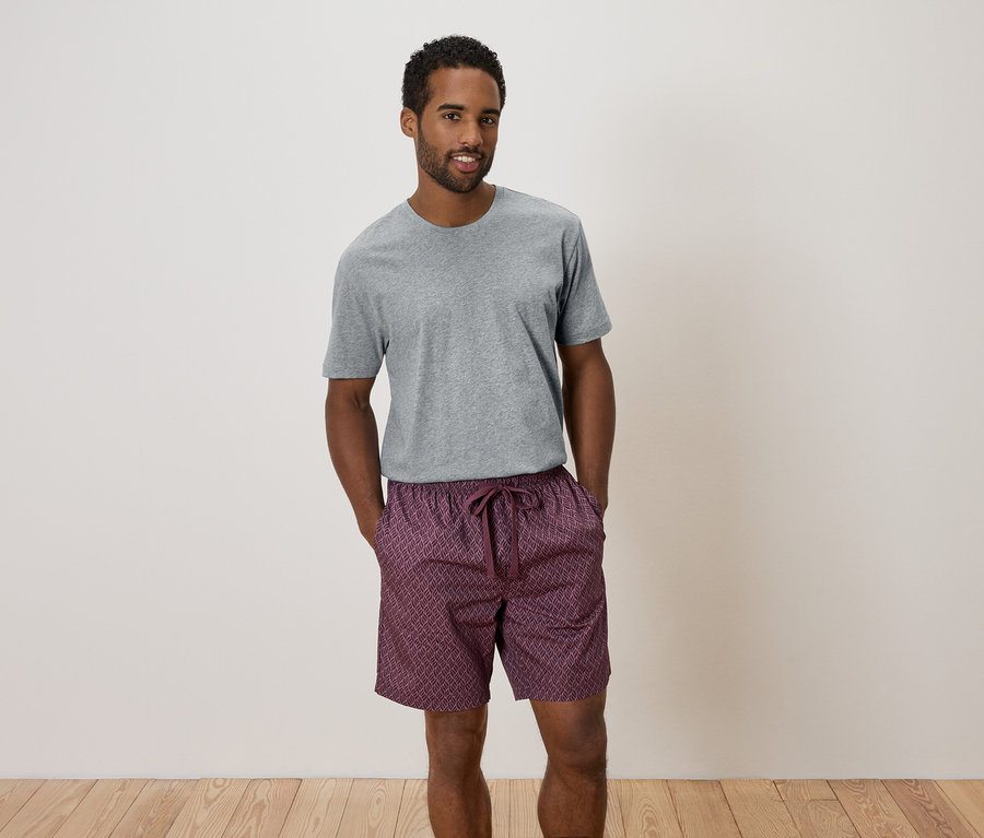 Un homme se tient debout, les mains dans les poches, portant un pantalon gris et un short à motifs.