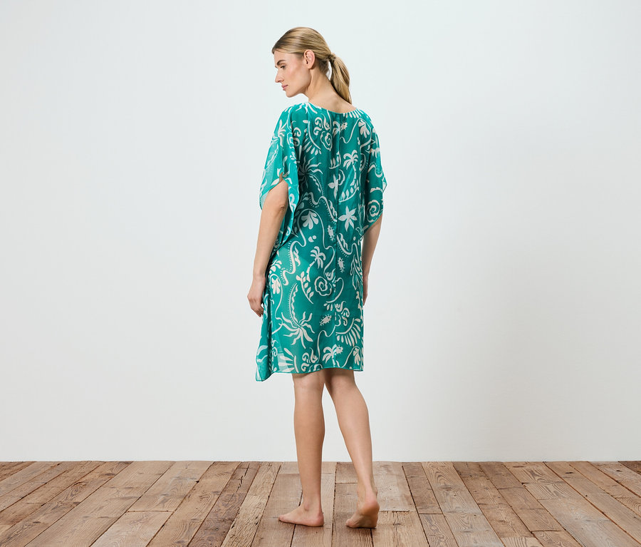 Une femme debout pieds nus porte une robe verte avec un motif blanc.