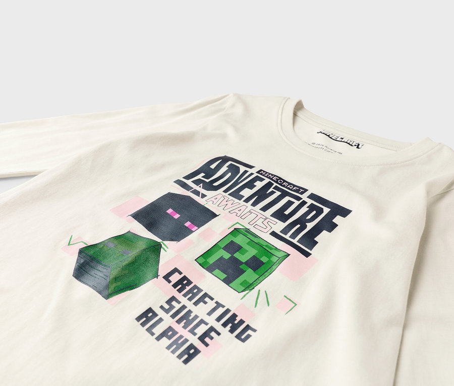 Sur une chemise blanche à manches longues figure un motif Minecraft avec l'inscription "Adventure Awaits" et "Crafting Since Alpha".