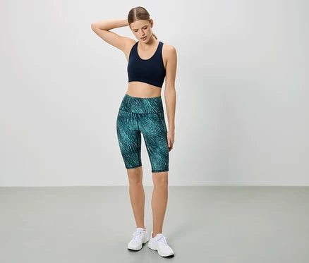 Une femme en tenue de sport pose devant un fond blanc. Elle porte un soutien-gorge de sport bleu marine et un legging court à motifs.