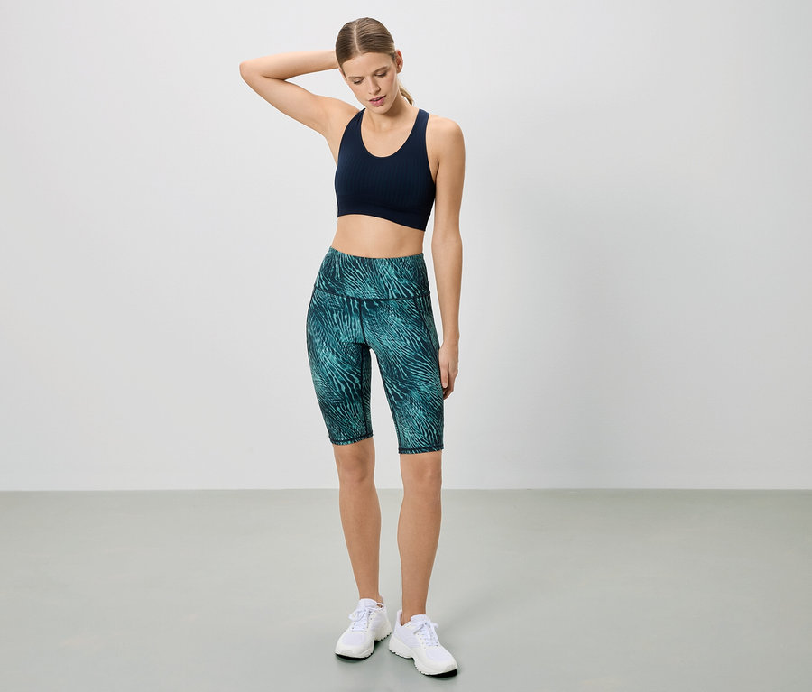 Une femme en tenue de sport pose devant un fond blanc. Elle porte un soutien-gorge de sport bleu marine et un legging court à motifs.
