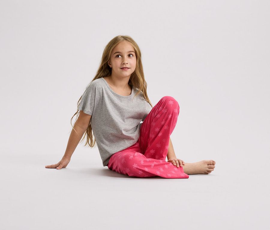 Une fille est assise par terre, portant un t-shirt gris et un pantalon rose avec un motif de palmiers.