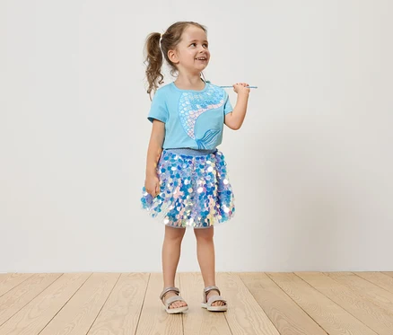 Fillette avec des couettes, portant un t-shirt bleu clair avec une sirène et une jupe à paillettes bleues, tenant une baguette magique.