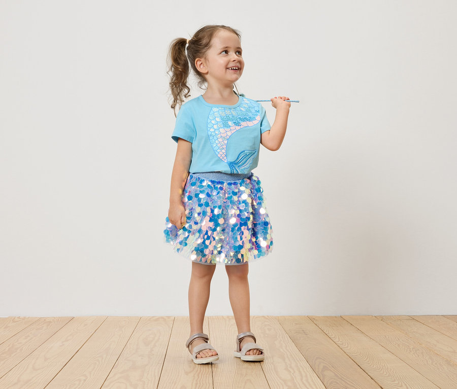 Fillette avec des couettes, portant un t-shirt bleu clair avec une sirène et une jupe à paillettes bleues, tenant une baguette magique.