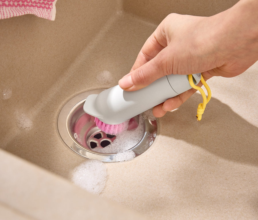 Une main nettoie le drain d'un évier beige avec une brosse grise à poils roses et une boucle jaune.