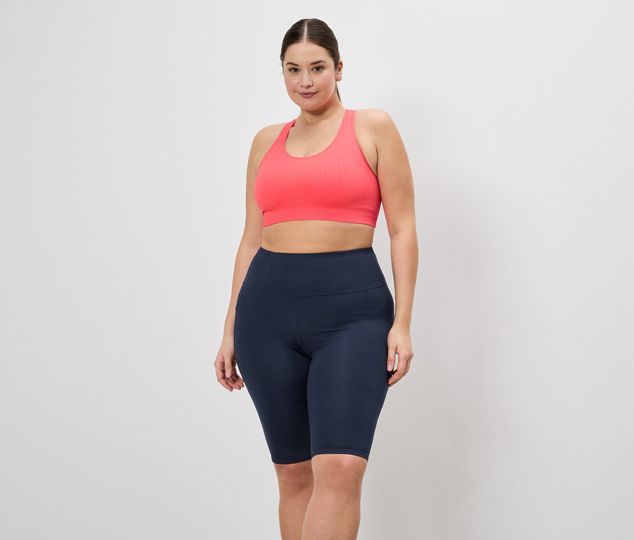 Une femme en tenue de sport pose devant un fond blanc. Elle porte une brassière de sport rose et un cycliste bleu marine.