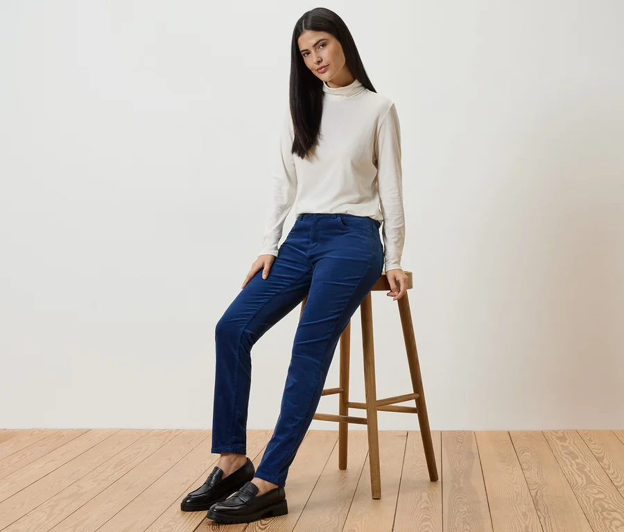 Une femme aux longs cheveux foncés est assise sur un tabouret et porte un pantalon en velours côtelé bleu, un haut crème et des chaussures noires.