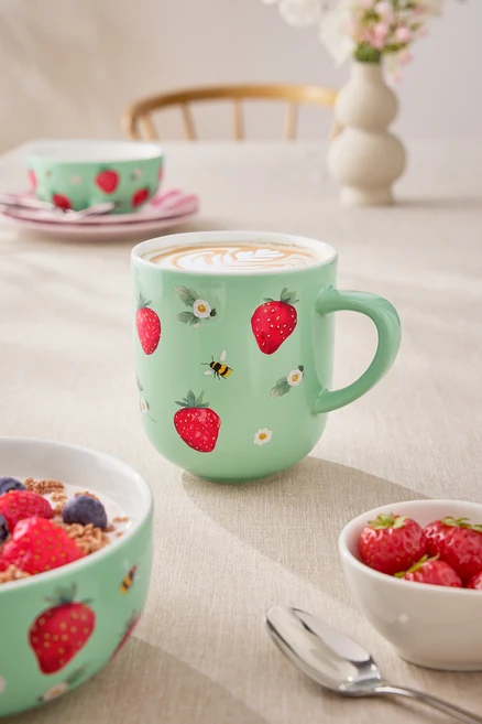 Gros plan d'une table avec une tasse de café vert clair, un bol de céréales avec du yaourt et des baies, et un petit bol de fraises. La tasse est décorée de fraises et de petites fleurs.