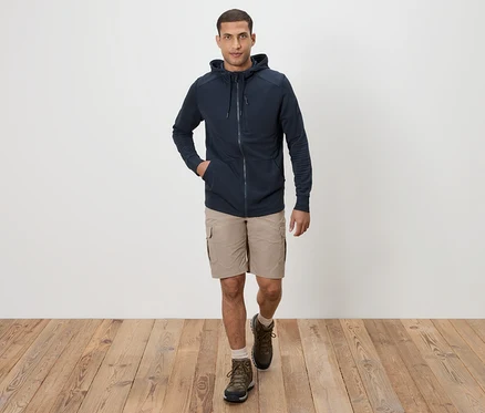 Un homme marche sur un plancher en bois en short kaki, un sweat à capuche bleu marine et des bottes marron.