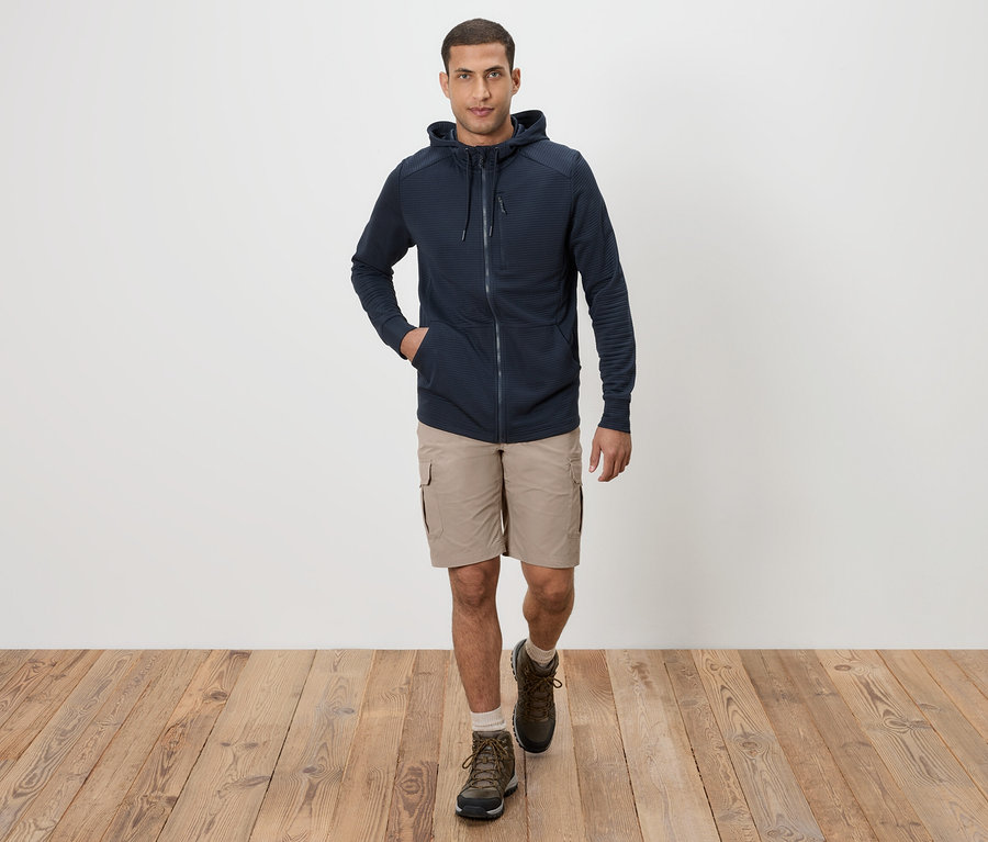 Un homme marche sur un plancher en bois en short kaki, un sweat à capuche bleu marine et des bottes marron.