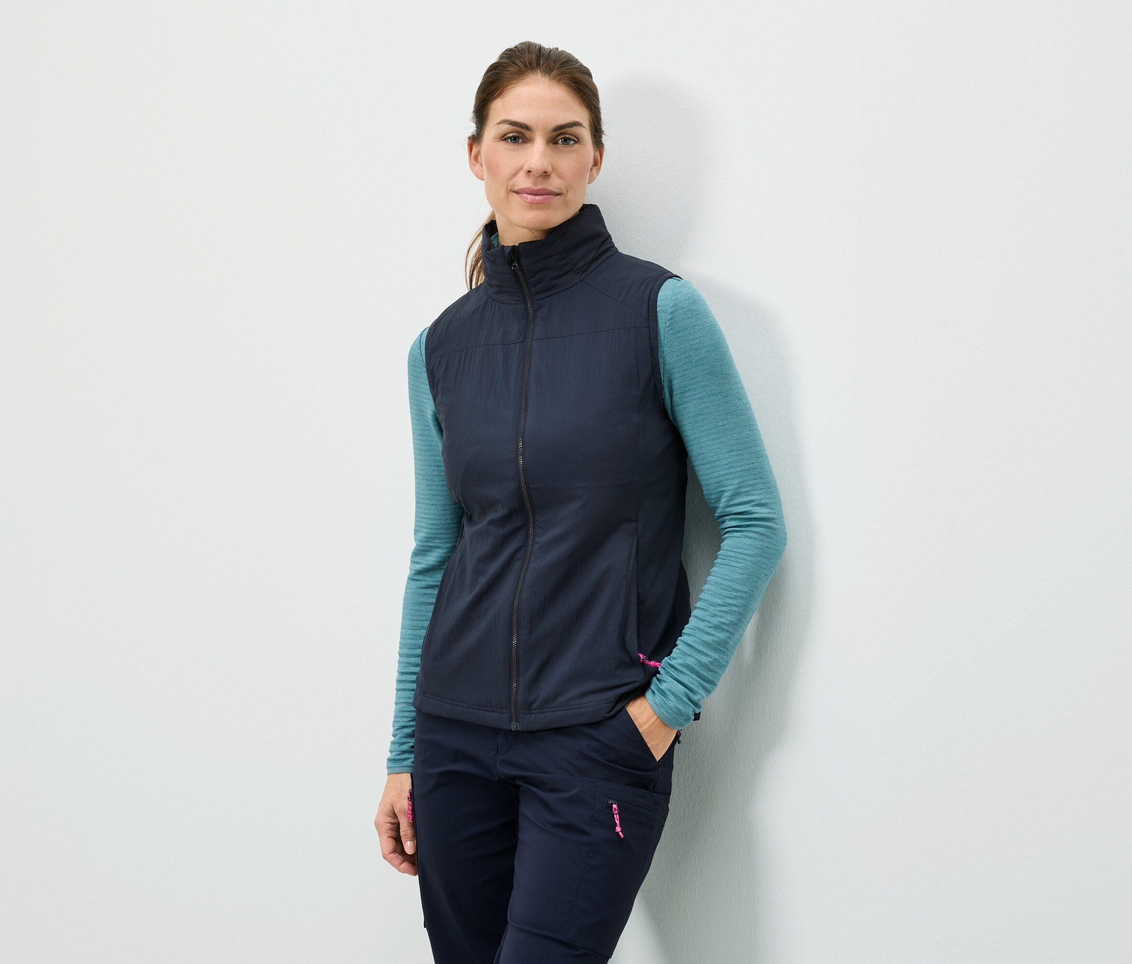 Une femme se tient adossée à un mur avec la main dans la poche, portant un gilet bleu marine, un haut à manches longues turquoise et un pantalon bleu marine.