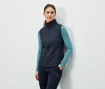 Une femme se tient adossée à un mur avec la main dans la poche, portant un gilet bleu marine, un haut à manches longues turquoise et un pantalon bleu marine.