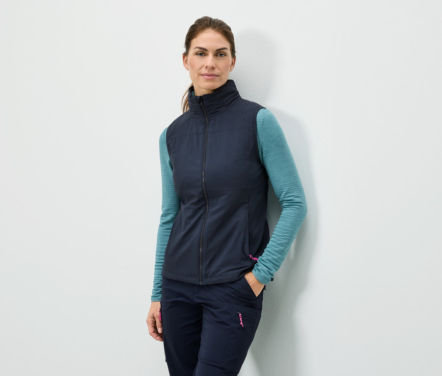 Une femme se tient adossée à un mur avec la main dans la poche, portant un gilet bleu marine, un haut à manches longues turquoise et un pantalon bleu marine.